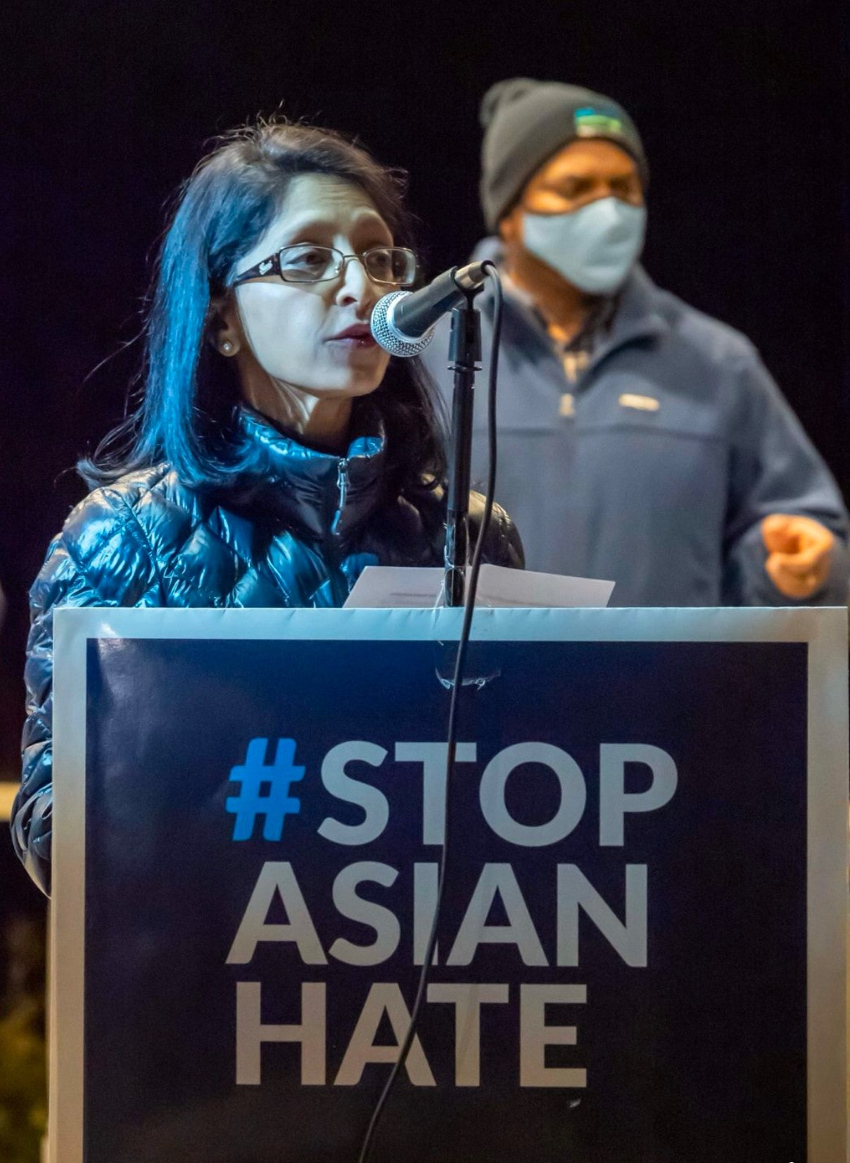 photo of vineeta in winter jacket speaking behind a microphone and a #stopasianhate poster at a night time event