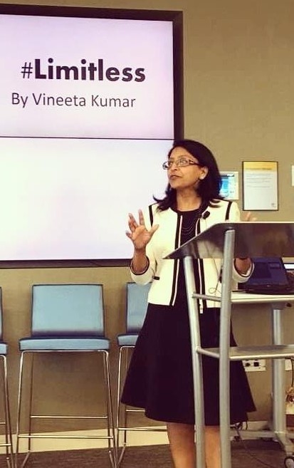 vineeta wearing a jacket and dress and speaking at a podium in front of a display that says #limitless by vineeta kumar