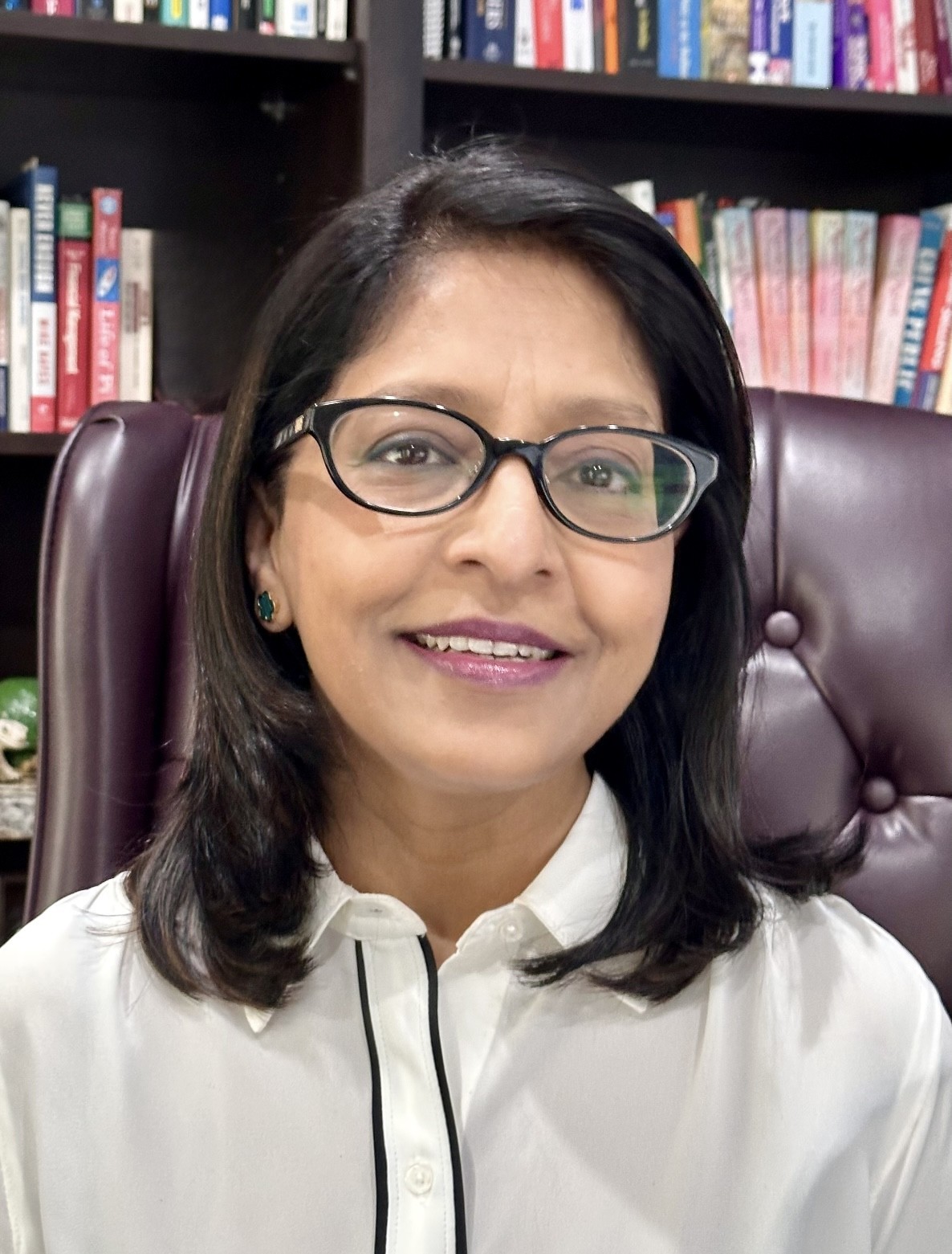 Headshot Vineeta Kumar, shoulder-length brown hair and glasses, smiling and sitting in leather office chair with shelves of library books in background.